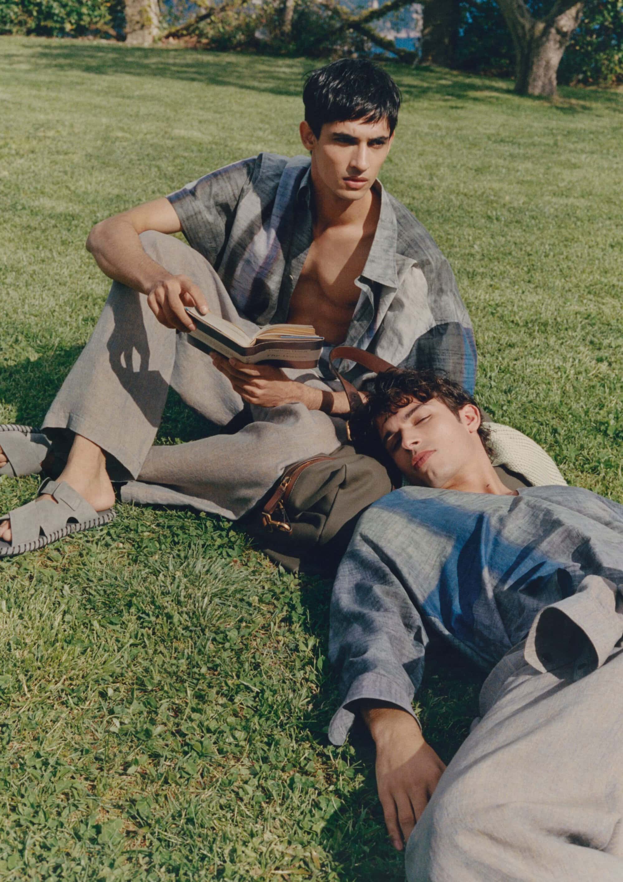 Two male models lounging on a sunlit grass lawn, one seated reading a book while the other rests lying down in loose shirts and grey trousers.