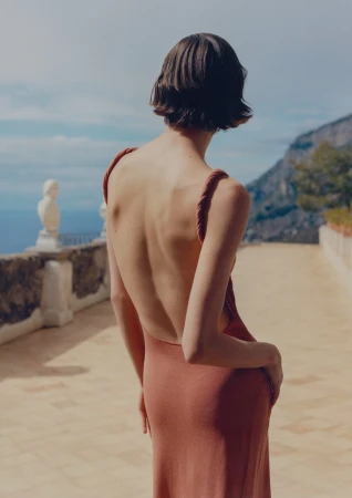 Woman with short brown hair wearing backless dress stands on stone balcony overlooking mountain range and ocean on sunny day.