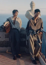 Two male models in relaxed neutral menswear pose beside a classical marble bust on a scenic cliffside terrace overlooking the ocean.