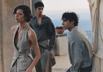 Model in a grey tailored halter vest posed outdoors on a stone terrace with two male models overlooking the sea.