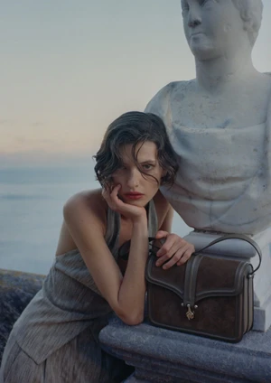 Valerie Scherzinger leaning beside a classical marble bust holding a brown suede Giorgio Armani handbag against a sea backdrop.