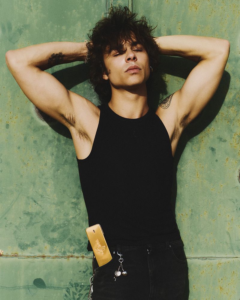 Young man with shaggy dark hair wearing a black tank top and jeans with a gold fragrance bottle in his pocket against a green wall.