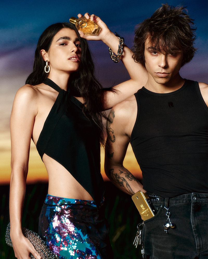 Two models in black designer tops holding Rabanne fragrance bottles against a dusk sunset sky.