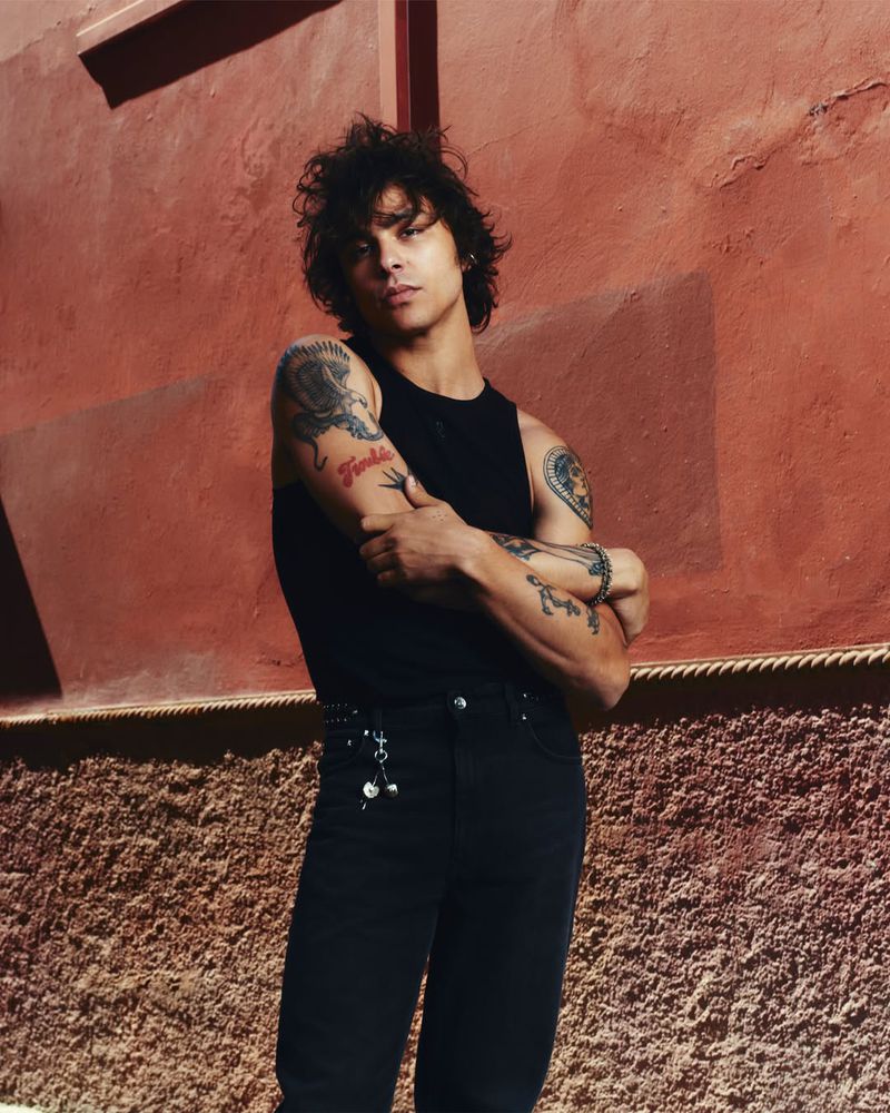 Portrait of a young adult man with curly hair and tattoos posing in black clothing against a textured orange wall in a studio setting.