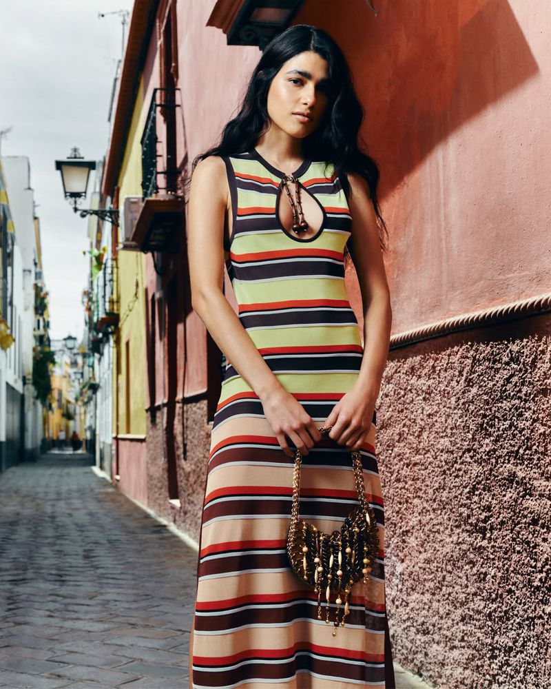Model Yarlin Mateo poses in a striped knit maxi dress with a keyhole neckline, holding a fringed handbag in a narrow alley.