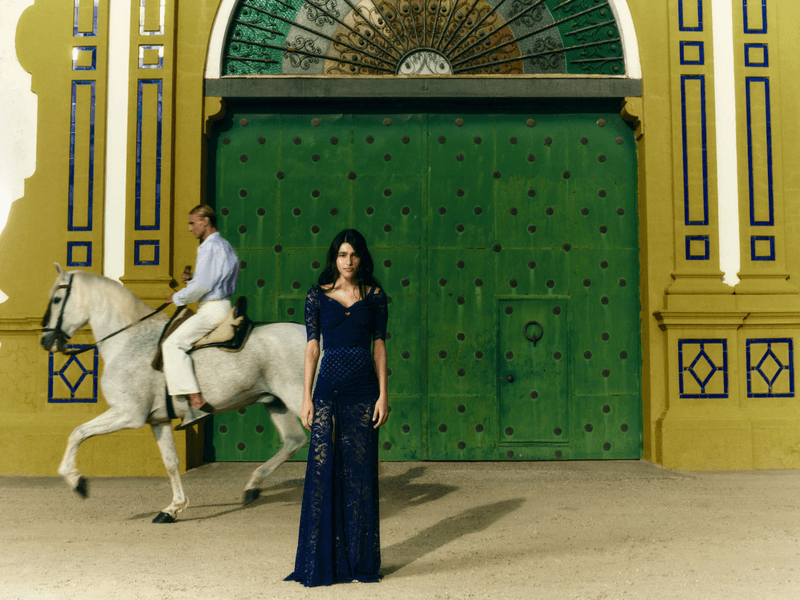 Model in a navy blue lace gown stands before a large green studded gate while a man on a white horse rides behind her.