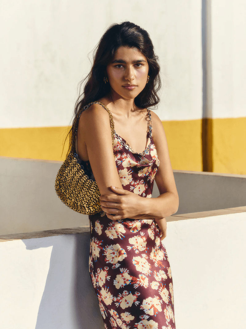 Model Yarlin Mateo wearing a burgundy floral slip dress and gold metallic shoulder bag in direct sunlight.
