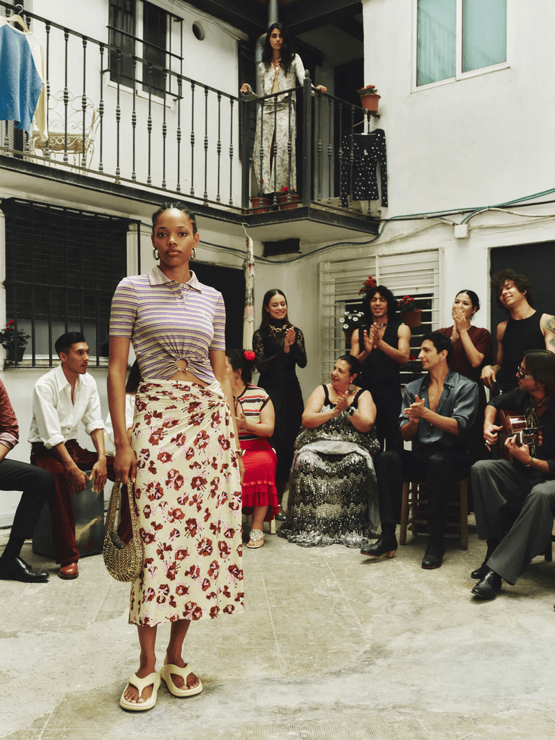 Model Yarlin Mateo wearing a striped crop top and floral midi skirt standing in a Spanish courtyard surrounded by clapping onlookers.