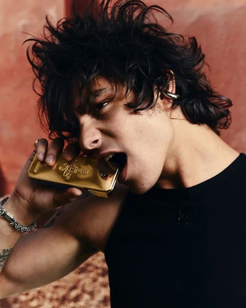 Yarlin Mateo wearing a black tank top and biting a gold Rabanne fragrance bottle against a warm textured wall.
