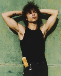 Young man with shaggy dark hair wearing a black tank top and jeans with a gold fragrance bottle in his pocket against a green wall.