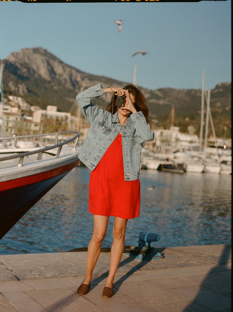 Constanze Saemann in a red mini dress and light blue denim jacket poses with a camera on a harbor dock.