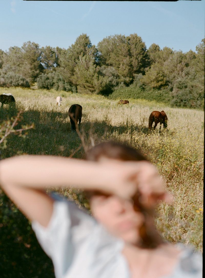 Model Constanze Saemann shielding her face in a sunlit meadow with grazing horses for Mavi
