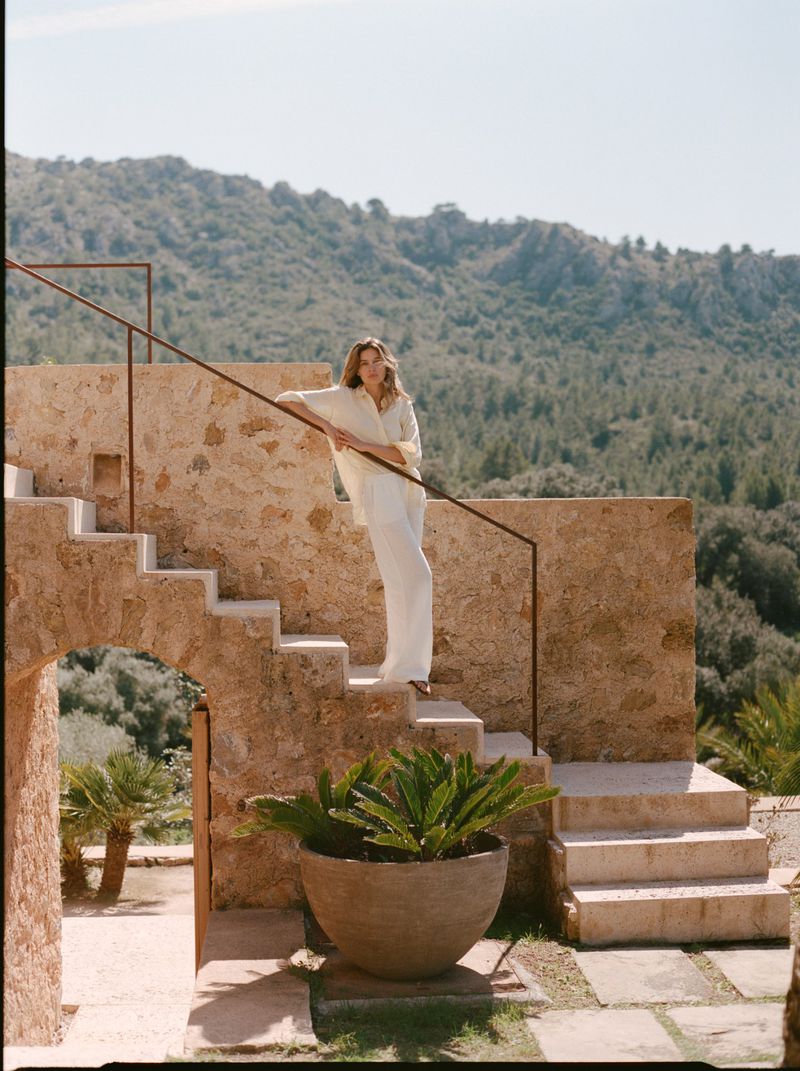 Model Constanze Saemann in a loose white button-down shirt and wide-leg trousers standing on rustic outdoor stone steps.