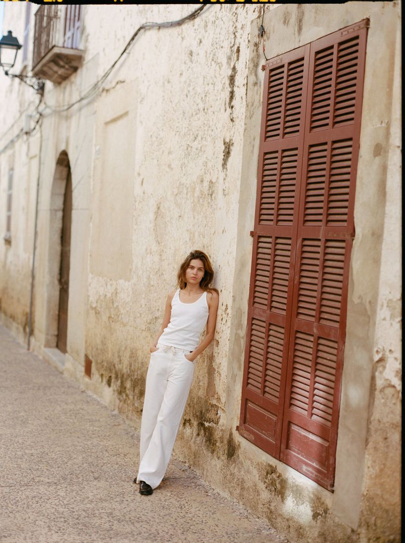 Model Constanze Saemann in a white tank top and white wide-leg trousers leaning against a textured building wall.