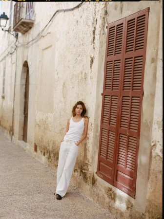 Model Constanze Saemann in a white tank top and white wide-leg trousers leaning against a textured building wall.