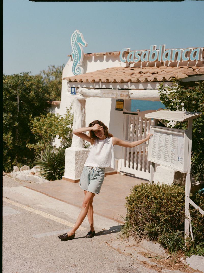 Model Constanze Saemann shields her eyes in a white sleeveless top and light blue shorts outside a coastal restaurant entrance.