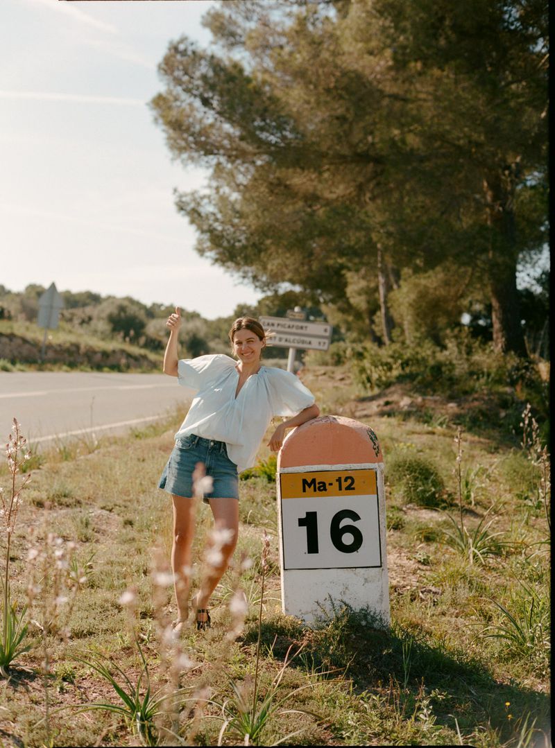Woman leans on a kilometer stone by a country road with trees and blue sky in the background, wearing a blouse and denim shorts.