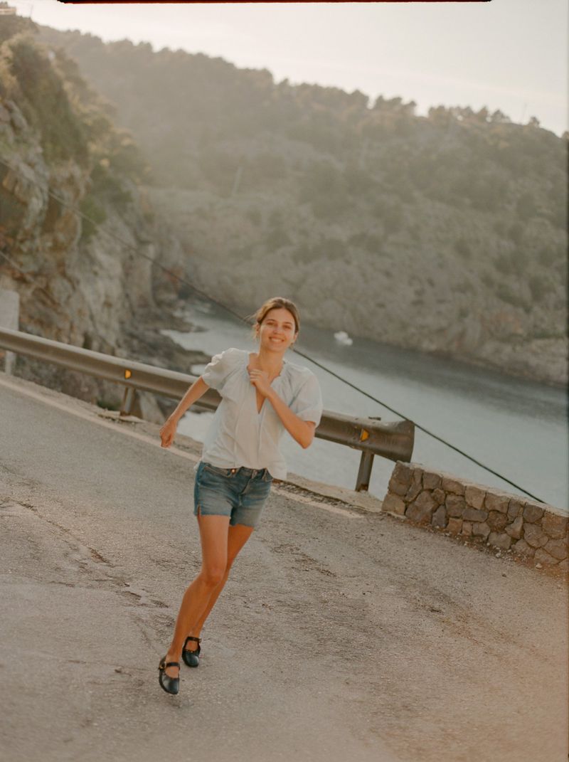 Constanze Saemann running down a coastal road wearing a light blouse, denim shorts, and black flats with a bay in the background.