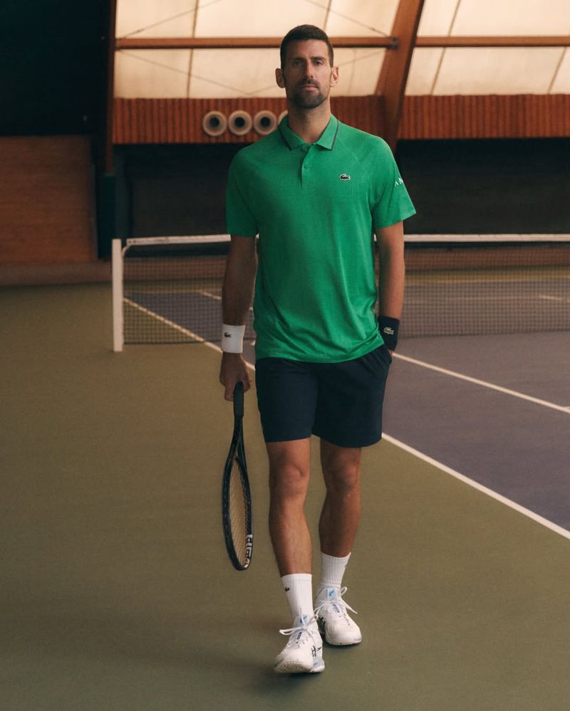 Professional male tennis player in green Lacoste polo shirt walking on indoor court, holding a racquet during match or practice.