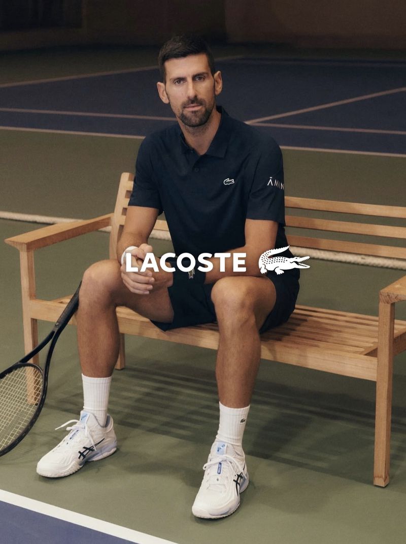 Novak Djokovic seated on a wooden bench on a tennis court holding a racket and wearing a navy Lacoste polo shirt.