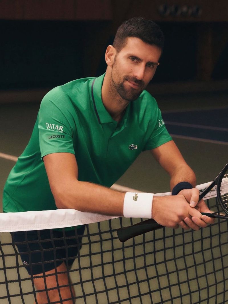 Novak Djokovic leaning on a tennis court net wearing a green Lacoste polo shirt and holding a tennis racket.