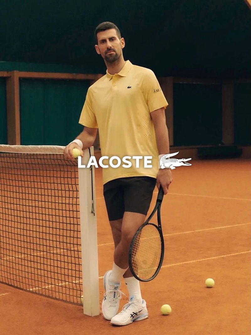 Novak Djokovic standing on a clay tennis court wearing a yellow polo shirt and black shorts holding a tennis racket