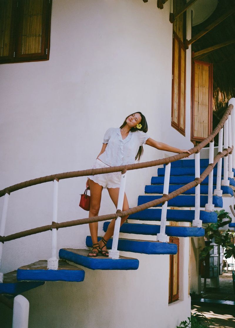 Didi Sviridova poses on an outdoor staircase with vibrant blue steps and rope railings against a white wall in light summer clothing.