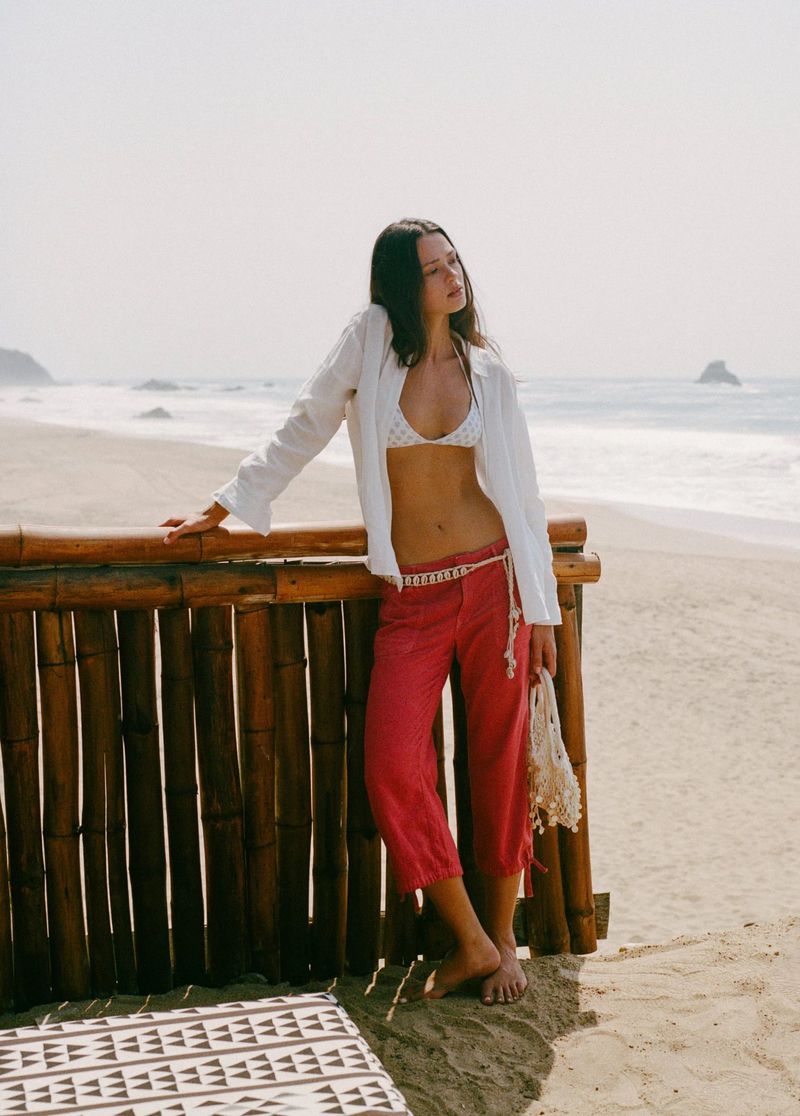 Model leaning against a bamboo fence on a sandy beach wearing a white shirt and red trousers, captured by Juan Hernández.