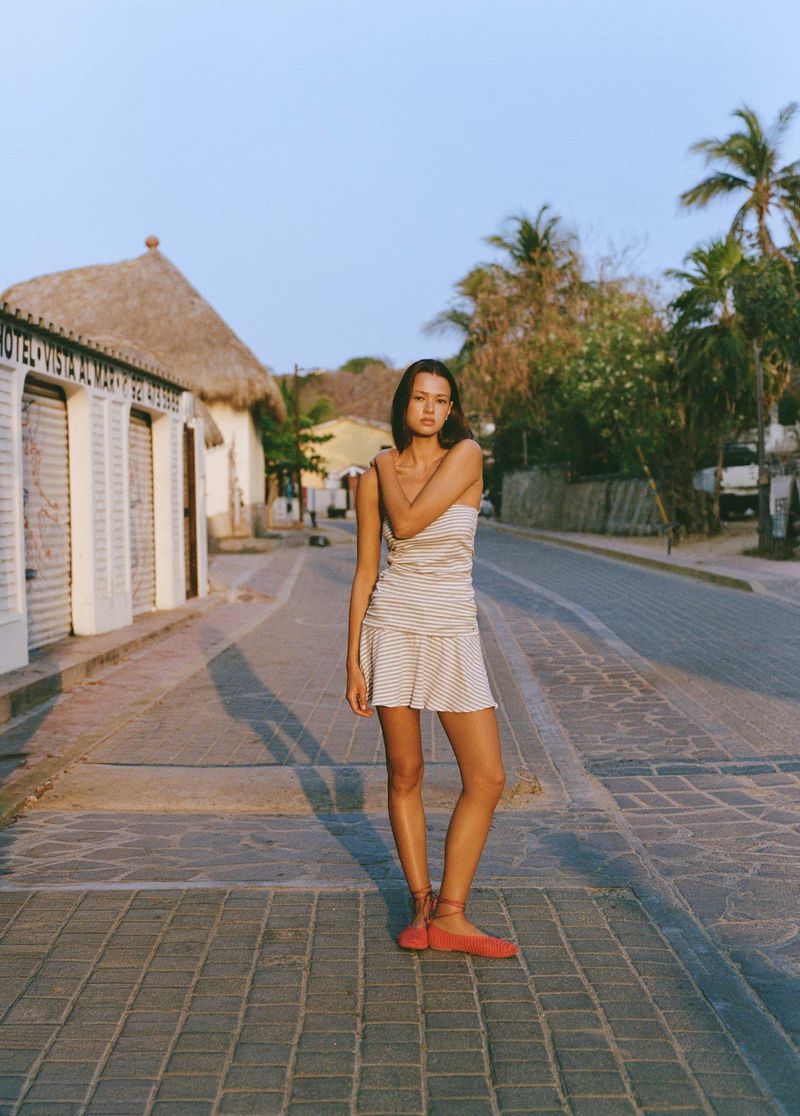 Model Didi Sviridova standing in a striped strapless mini dress and red lace-up flats on a paved village street.