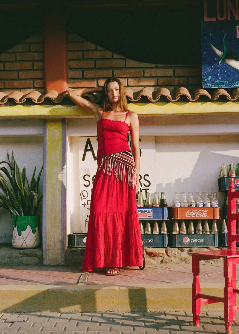 Model Didi Sviridova in a red maxi dress and crochet fringe belt standing in front of stacked soda crates outdoors.