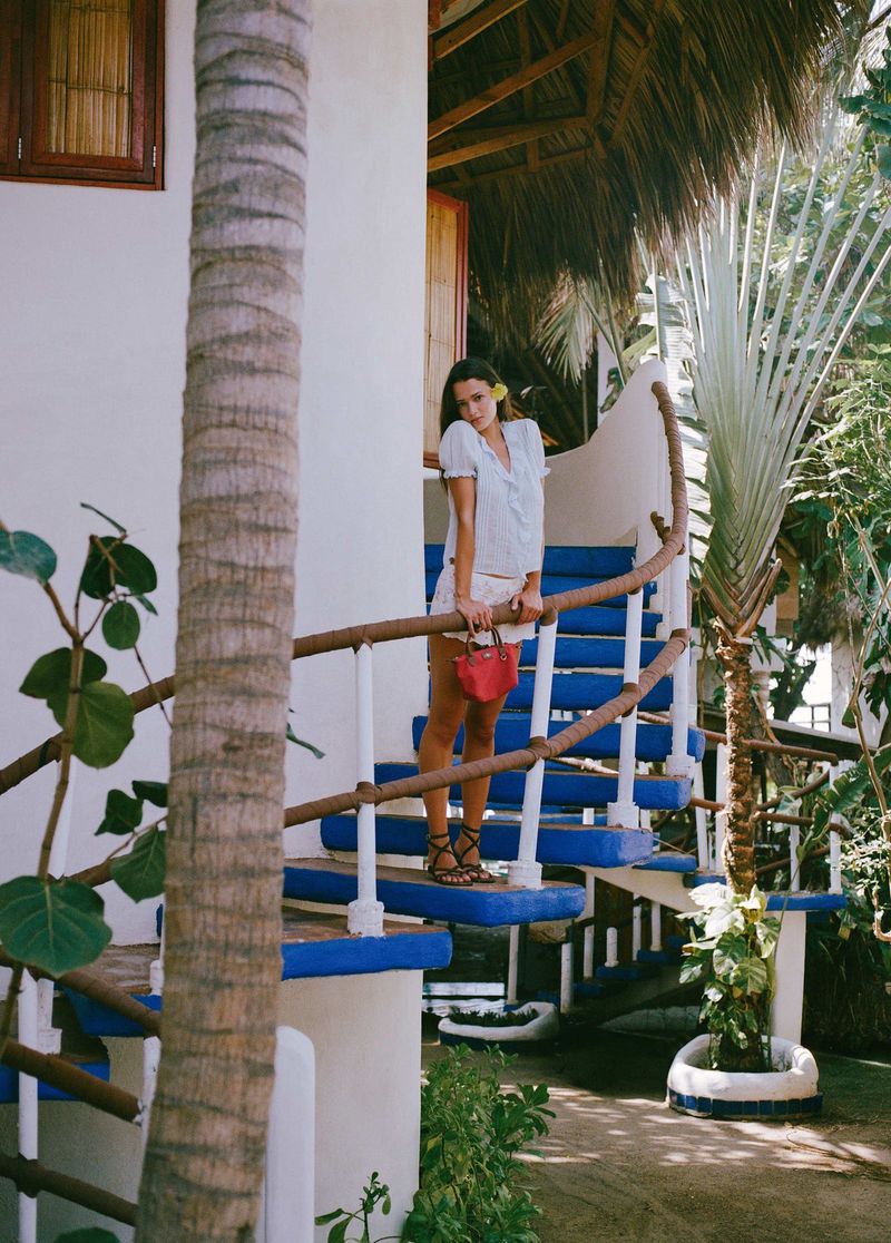 Didi Sviridova in a white summer outfit holding a small red handbag on vibrant blue and white stairs surrounded by palm trees.