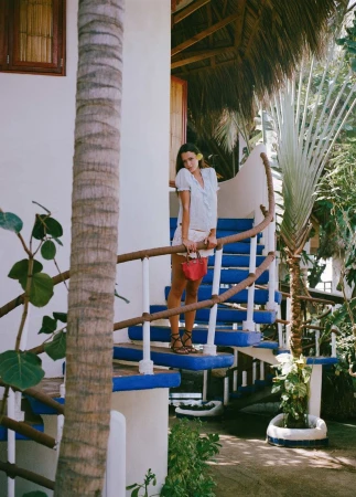 Didi Sviridova in a white summer outfit holding a small red handbag on vibrant blue and white stairs surrounded by palm trees.