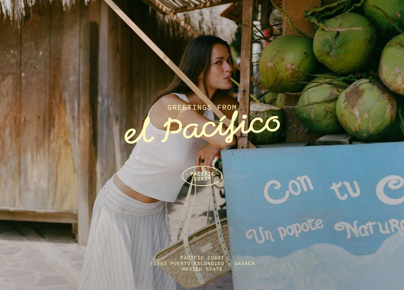 Model Didi Sviridova leaning by a rustic green coconut stand holding a woven tote bag.