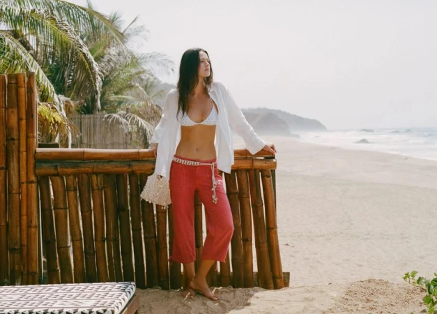 Model Didi Sviridova standing by a bamboo fence on a sandy beach wearing a white shirt and coral cropped trousers.