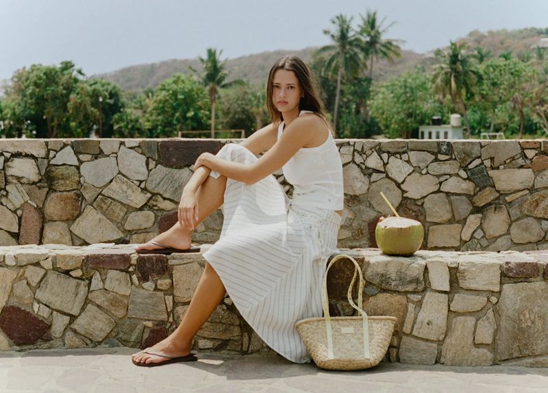 Model Didi Sviridova sitting on a stone wall in a white tank top and striped skirt next to a straw tote bag and coconut.