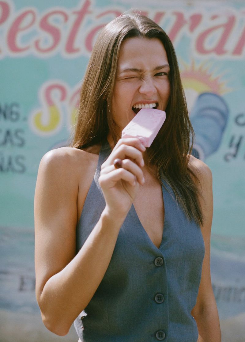 Model Didi Sviridova wearing a blue halter vest and winking playfully while biting a pink popsicle outdoors.