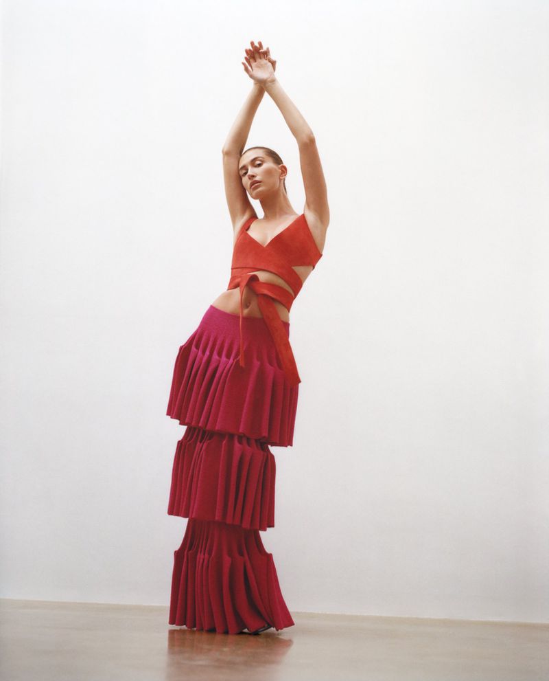 Hailey Bieber posing in a red wrap crop top and a tiered pleated crimson maxi skirt against a white studio background.