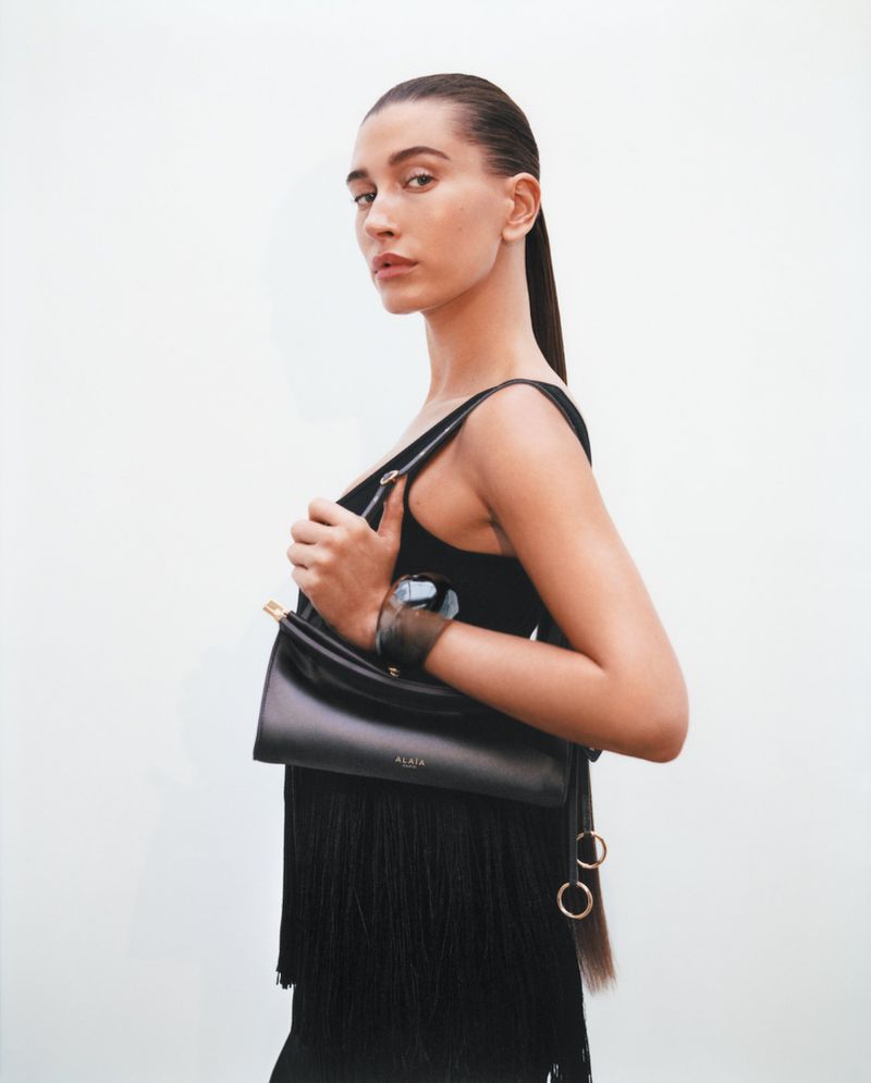 Hailey Bieber holding a black leather Alaïa handbag and wearing a black fringe top against a solid white studio background.