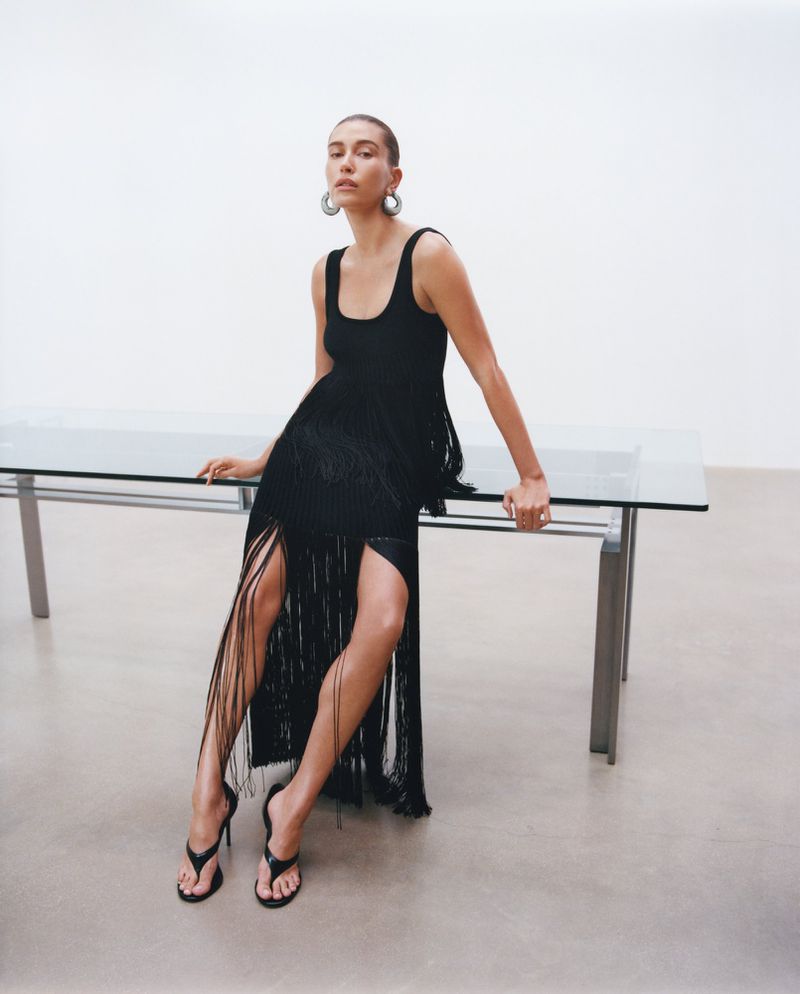 Hailey Bieber wearing a black fringe dress and heeled sandals while leaning against a glass table for an Alaïa fashion campaign.