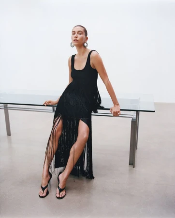 Hailey Bieber wearing a black fringe dress and heeled sandals while leaning against a glass table for an Alaïa fashion campaign.