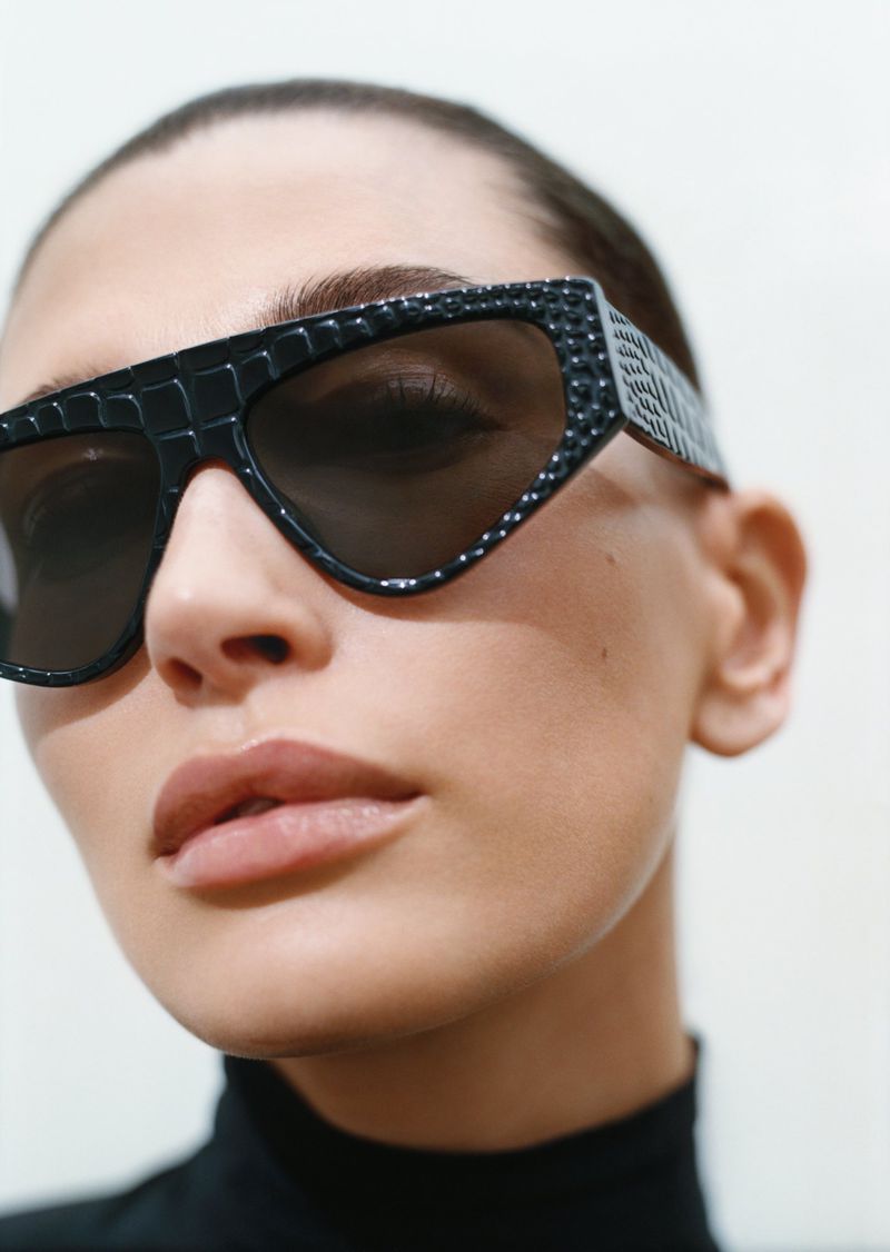 Hailey Bieber wearing textured black geometric sunglasses and a black turtleneck against a plain white studio background.