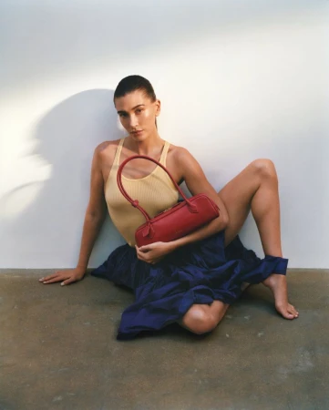 Hailey Bieber seated on the floor in a pale yellow ribbed tank top and navy blue skirt holding a red leather handbag.