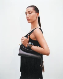 Hailey Bieber holding a black leather Alaïa handbag and wearing a black fringe top against a solid white studio background.