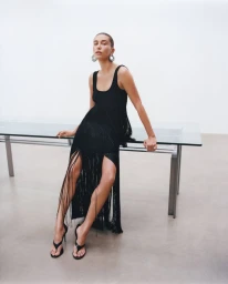Hailey Bieber wearing a black fringe dress and heeled sandals while leaning against a glass table for an Alaïa fashion campaign.