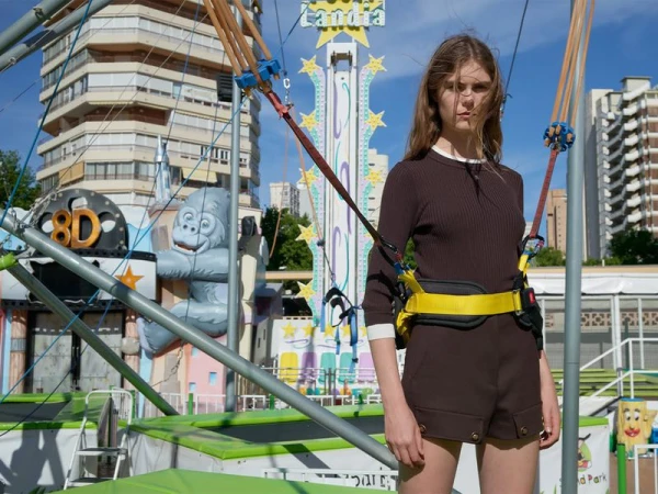 A slender woman in brown shorts and matching top stands in front of a trampoline park, high-rise buildings in the background.