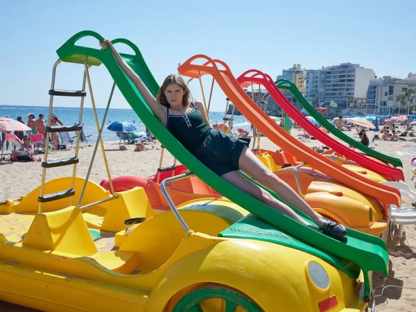 A woman is sliding on a green slide on the beach next to colorful slides, people, buildings and the blue sea under a bright sunny day.