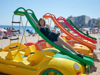 Model reclining on a colourful paddle boat slide on a sandy beach under bright daylight with ocean and buildings in background.