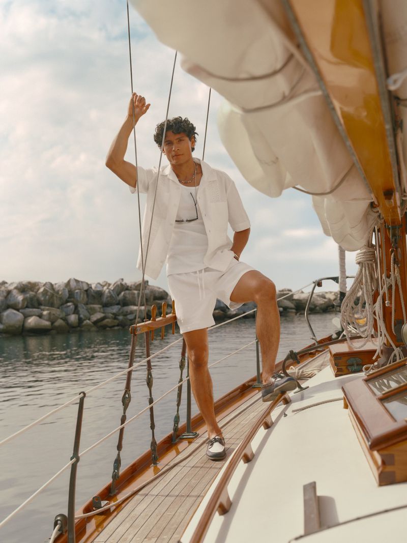 Model Hitesh Bisht in an all-white short-sleeve shirt and shorts standing on the wooden deck of a sailboat near a rocky shoreline.