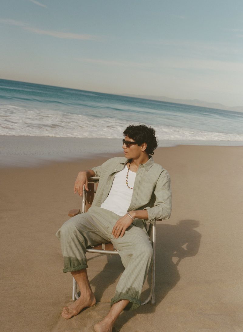 Man in a sage green shirt and trousers sitting on a folding chair on a sandy beach facing the ocean waves.