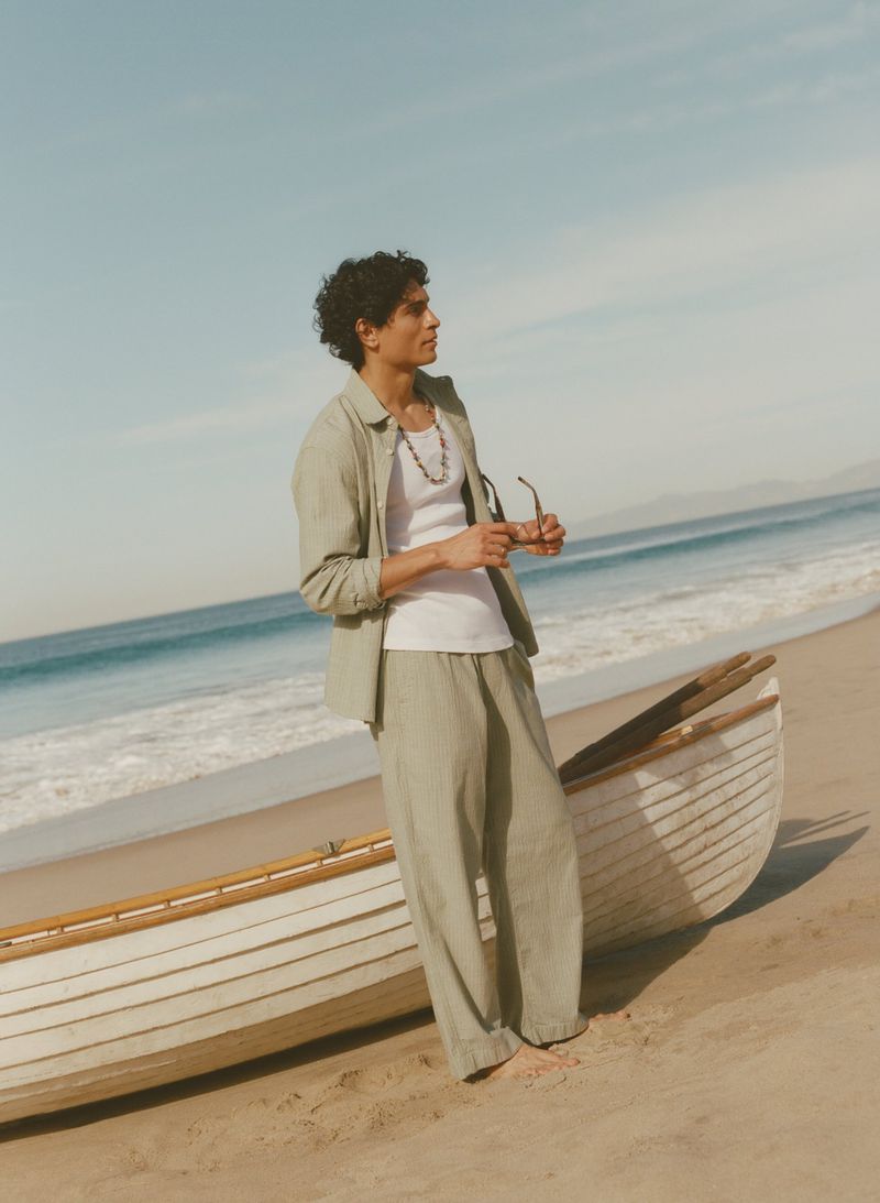 Man in a light green linen shirt and trousers standing barefoot beside a wooden rowboat on a sandy beach.